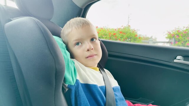 A young boy rests comfortably in a car seat. He gazes thoughtfully during a peaceful ride past bright flowers outside.