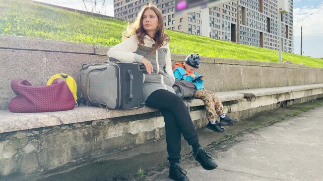 A mother and son sit with luggage on a street in Saint Petersburg. The woman looks away pensively while her boy plays calmly on a phone. They feel tired yet patient during travel.