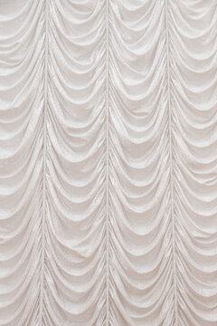 Cream fabric curtain features repeated scalloped swags and vertical pleats across the full surface, forming a soft ornamental backdrop with layered folds and neutral tones in an indoor hall