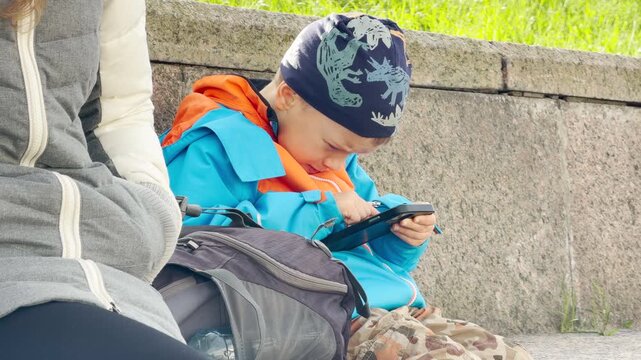 A young boy interacts eagerly with a smartphone. He feels happy and focused while resting on a granite embankment in Saint Petersburg.