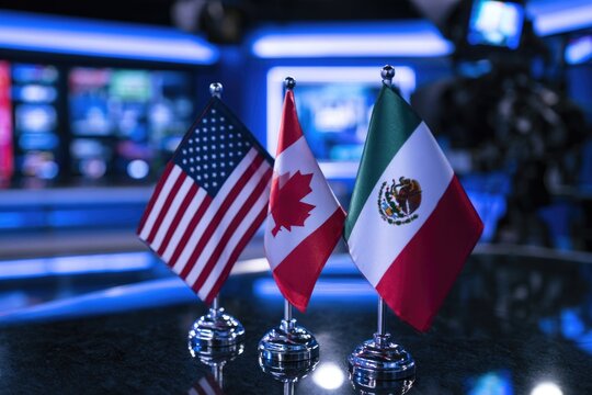 Three small flags of the United States, Canada, and Mexico stand side by side on a reflective surface with a blurred modern background.