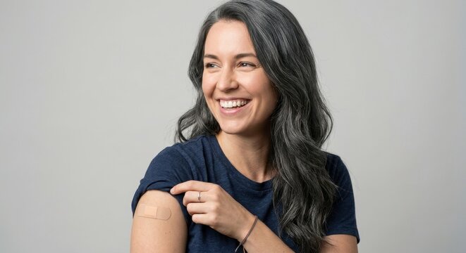 Happy mature woman with grey hair smiling and showing an adhesive bandage on her shoulder after getting a vaccine shot or immunization at a clinic, healthcare and disease prevention concept.