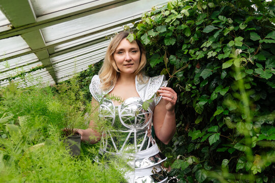 A woman stands inside a greenhouse wearing a shiny silver outfit