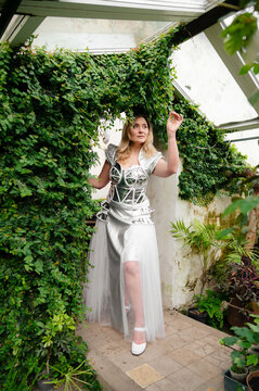 Model poses in a unique outfit inside a greenhouse surrounded by plants during the day