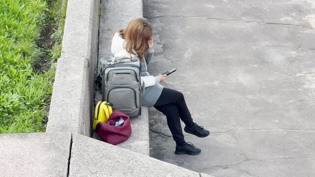A woman traveler sits with her luggage and checks her phone on a stone ledge in Saint Petersburg. She feels calm and patient while waiting. The peaceful scene brings a gentle mood.