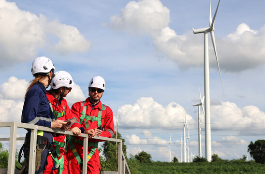 A team of professional engineers, both men and women, in red and navy safety uniforms, are discussing maintenance at a renewable energy wind farm that generates electricity. 