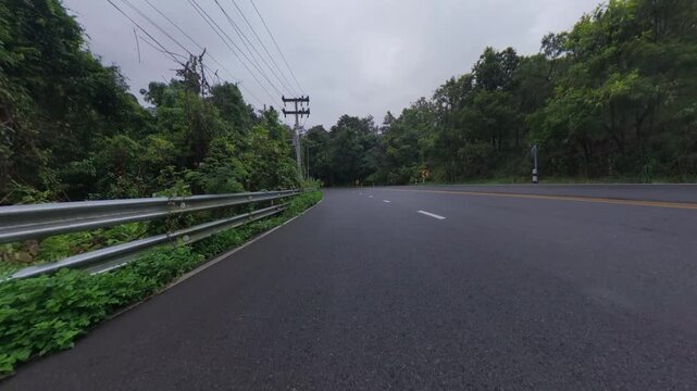 Cyclist POV footage riding on a scenic asphalt road through lush green forest, with guardrails, utility poles and tropical vegetation under an overcast sky.