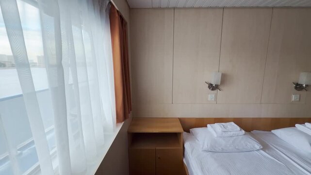 A neat bed rests inside a bright cruise ship cabin on the Neva River. Sheer curtains sway gently as soft daylight filters into the cozy room. The quiet space offers a peaceful and serene feeling.