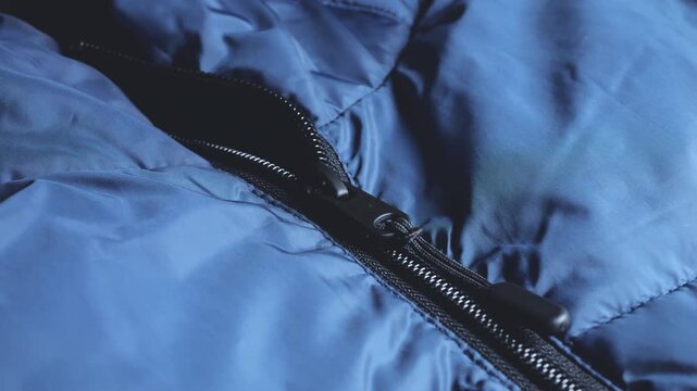 A close-up of a blue padded jacket with a black zipper puller shows dark navy outerwear, quilted waterproof fabric texture, functional hardware, large plastic fastener tracks and chilly autumn weather