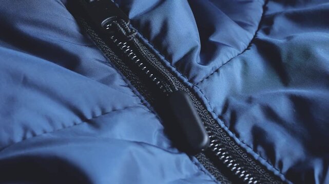A close-up of a black zipper puller on a blue puffer jacket shows outerwear on a flat surface, the texture of quilted waterproof clothing fabric, and plastic zipper tracks.