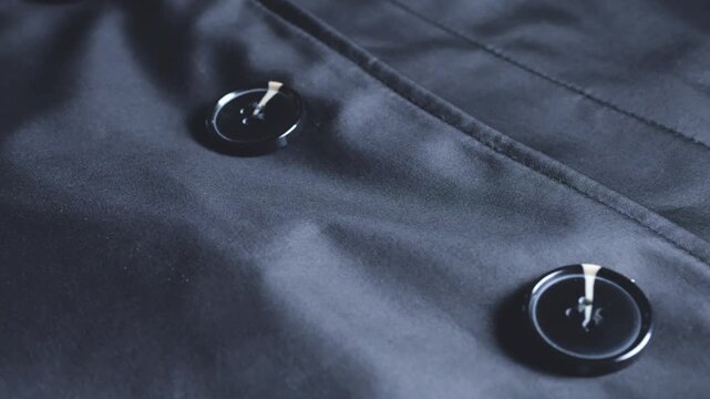A close-up of a black trench coat fabric with buttons shows the classic dark outerwear texture, high-quality sewing elements, professional tailoring, and an elegant, practical style.