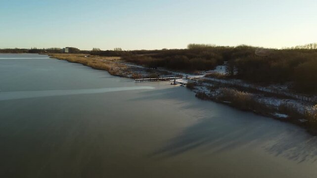 Frozen Ega Engso lake wetland and snow-dusted shoreline near Aarhus on a clear winter day, aerial orbit of wooden dock with sunset golden glow