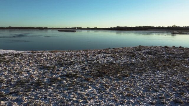 Snowy shore meets frozen Ega Engso lake wetland under clear blue winter sky near Aarhus, Denmark, aerial tracking right along ice covered area