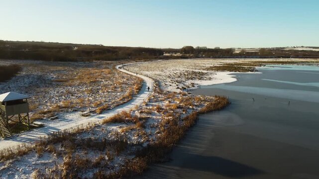 Frozen Ega Engso lake wetland beside winding snowy trail and winter reeds near Aarhus, Denmark, aerial medium orbit from viewing platform