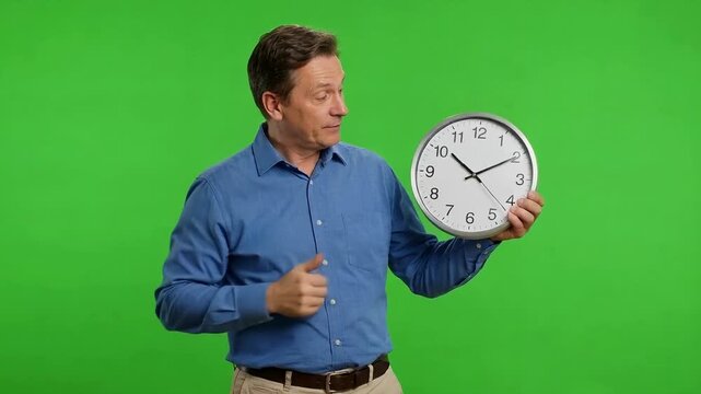Middle-aged man in a blue shirt points to a large analog clock against a bright green chroma key background