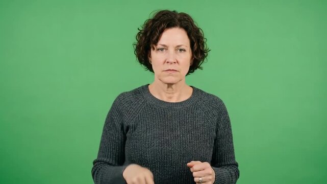 A serious curly-haired woman in a dark sweater points downward while communicating with expressive hand gestures against a solid green background