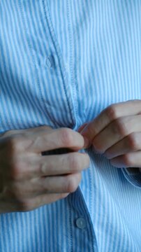 Woman hands fastening buttons on light blue and white striped cotton shirt, close up view of getting dressed in morning or preparing for work and starting new day. Vertical video