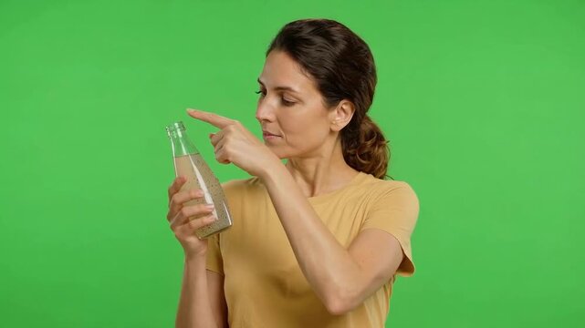 Young woman in a mustard shirt examines and points at a clear glass bottle against a bright green chroma key background