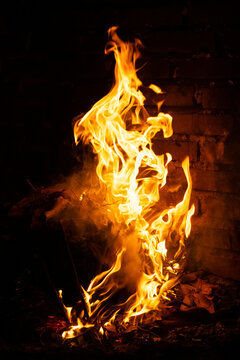 Bright orange and yellow flames blazing against a dark background. The flames originate from a small fire or burning wood.