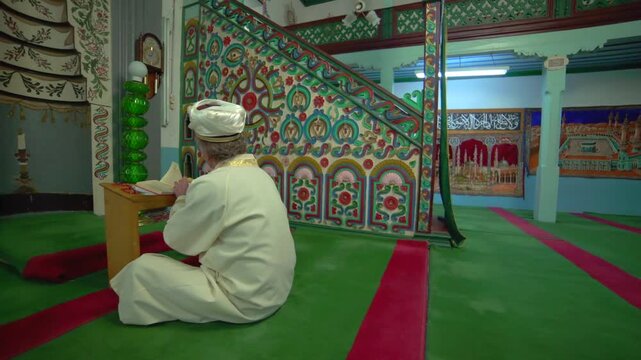 Historic wooden mosque interior with imam, worshippers, carpets, and Islamic prayer setting.
