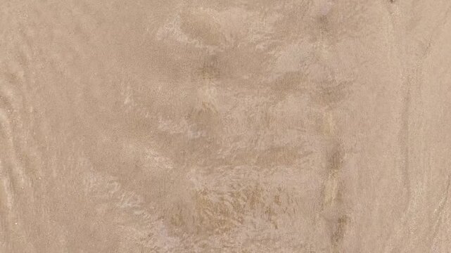 Close-up footage of shallow clear water gently flowing over a sandy riverbed, with sunlight creating rippling caustic patterns across the textured sand surface in a calm natural setting.