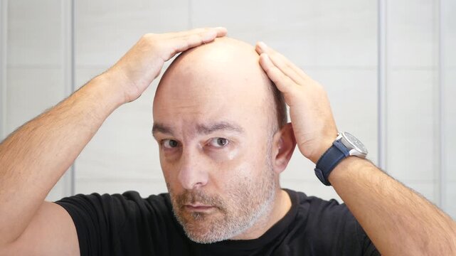 Bald middle aged man examines his head in the mirror during a private self image moment.