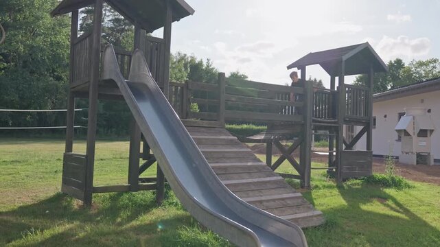 A boy on playground sliding down slow motion a metal slide at a wooden playground