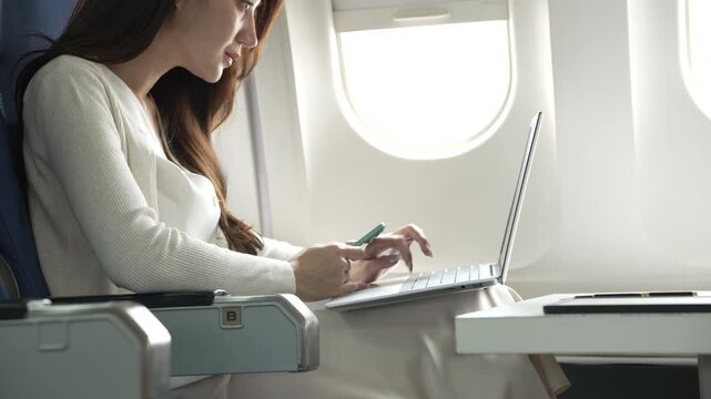 Young woman using a laptop and holding a card while seated by an airplane window, working during a flight in a bright modern cabin.