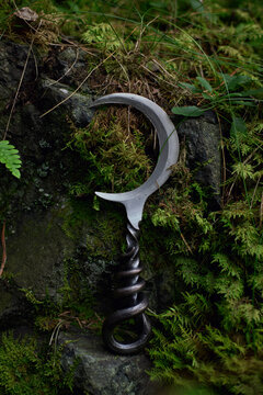 Single crescent blade rests vertically on a mossy rock face beneath fine grass and leaves in a woodland still life photograph with a dark twisted handle and bright metal edge