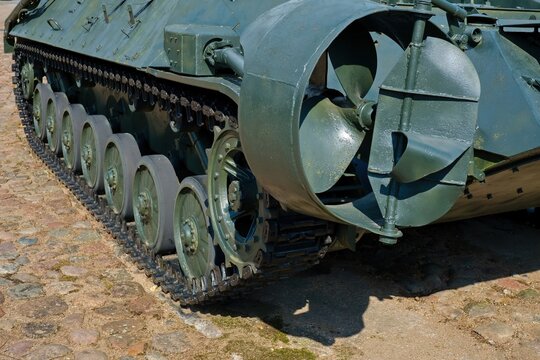 Close-up of caterpillar tracks, road wheels and water propeller shroud of a vintage Soviet IRM Zhuk amphibious armored engineering reconnaissance vehicle at an outdoor military exhibition.