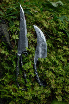 Paired steel knives with long pointed and broad curved blades rest against lush forest moss in a close vertical still life with twisted dark handles and contrasting silver textures