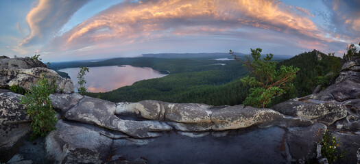 Curved rock ledge overlooks a forest lake reflecting pink sunset clouds in a panoramic landscape photograph with a pine tree at the edge for peaceful travel themes © angel_nt