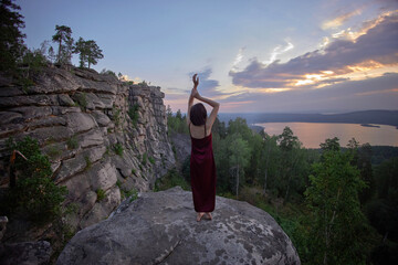 Woman in a burgundy dress raises her arms above a rocky lake overlook in a rear view sunset photograph with forest and distant water for freedom and reflection themes © angel_nt