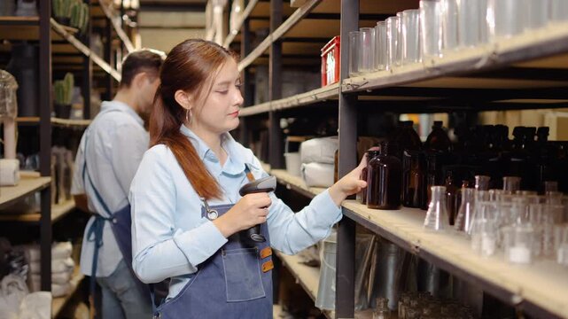 Asian female small business owner in a denim apron uses a handheld barcode scanner to check the inventory of glass bottles on warehouse shelves, while a male colleague organizes stock nearby.