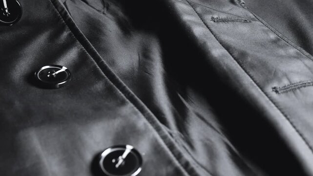 A close-up of a black satin trench coat with large round buttons reveals smooth waterproof fabric, neat seams, elegant classic outerwear details, and shiny elements on high-quality synthetic material.