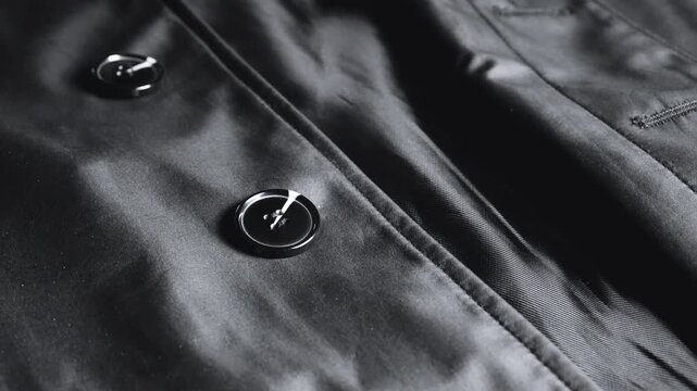A close-up of a black satin trench coat with large round buttons reveals smooth waterproof fabric, neat seams, elegant classic outerwear details, and shiny elements on high-quality synthetic material.