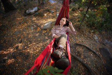 Woman reclines in a red striped hammock among woodland trees in a sunlit outdoor photograph with hiking boots and fallen leaves for camping relaxation and leisure themes © angel_nt