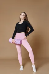 Woman in a black crop top and pink leggings holds a lavender foam roller in a full length studio fitness photograph on a tan background for exercise and recovery themes © angel_nt