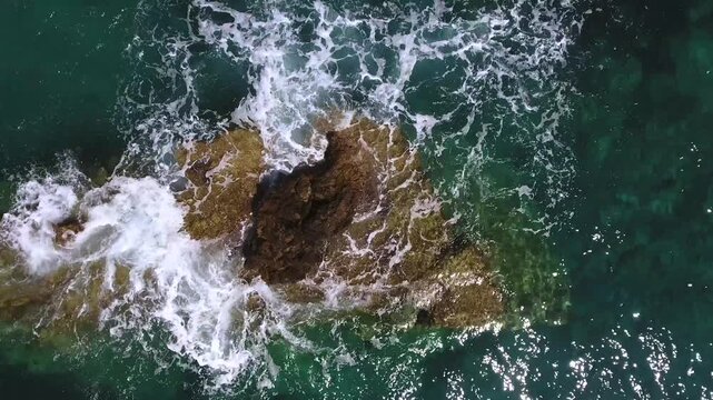 Aerial rock islet in turquoise sea with waves crashing over reef and foamy coastal water.