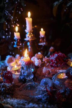 Glowing candles and melted wax stand among flowers and glass holders in a dark fantasy tabletop photograph with smoke and blue accents for atmospheric ritual decoration