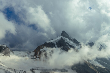 Triangular snowy mountain peak is partly wrapped in billowing white cloud in a landscape photograph with icy slopes and blue sky openings for alpine wilderness travel themes © angel_nt