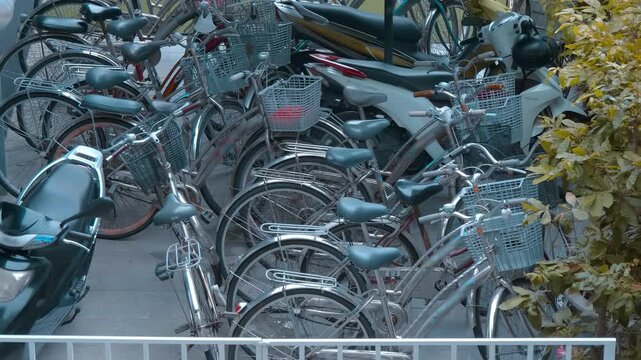 A crowded parking area filled with multiple commuter-style step-through bicycles (resembling Japanese mamachari utility bikes) equipped with front baskets and light-colored saddles, alongside a parked