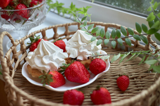 Cream topped pastries and red strawberries fill a white plate in a wicker tray near a rainy window in a close food photograph for cozy dessert and afternoon tea themes