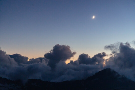 Slender crescent moon hangs above dark clouds and distant mountain silhouettes in a wide twilight landscape photograph with a peach horizon for quiet evening sky backgrounds