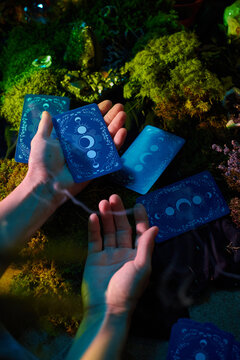 Hands handle blue cards with moon phase designs above a dark mossy tabletop in a vertical fantasy photograph with colored light and smoke for divination and mystery themes
