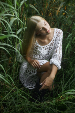 A young woman sits among tall grasses wearing a white dress. She has long hair and appears to be lost in thought. This scene takes place in a natural setting during daylight