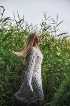 A woman enjoys the outdoors in a field surrounded by tall grass. She has long hair and wears a white dress. Sunlight shines through the leaves above her