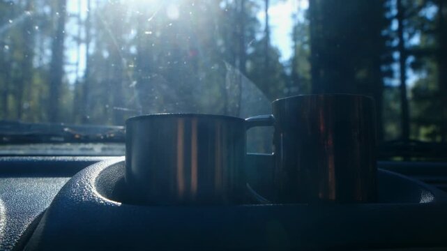 Two metal mugs with a steaming hot beverage resting on a car dashboard during an early morning stop in a sunny forest, creating a cozy and adventurous atmosphere for a road trip breakfast