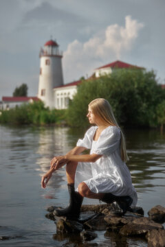 A young woman sits on rocks at the edge of a body of water. She wears a white dress and black boots. A lighthouse stands in the background with green bushes nearby. The sky is clear