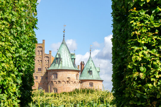 Egeskov Castle&rsquo;s red brick Renaissance facade and green copper spires peek above tall manicured hedges beside its reflective moat on a clear sunny day in Denmark